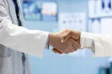 Twee personen in witte jassen schudden elkaar de hand, wat een professionele samenwerking of overeenkomst suggereert.